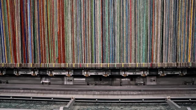 Warp threads pass in order through a carpet weaving machine on a production line. This video frame shows carpet manufacturing, yarn alignment, textile process, and industrial machine detail.
