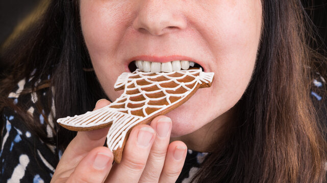 Closeup of a female face with gingerbread fish in mouth at eating Christmas pastry. Woman with long brown hair when is holding in hand and biting into sweet baked Xmas cookie decorated by white icing.
