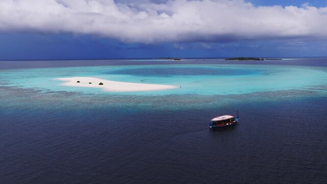 Aerial view of Dhigurah Island, Maldives, with crescent sandbank, three shrubs, lagoon rings, coral reef, distant islets, and a dhoni moving with faint wake.