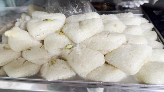 Close-up 4K footage of creamy Indian milk barfi squares topped with almonds and pistachios, neatly stacked behind a glass counter in a traditional sweet shop, highlighting festive dessert texture.
