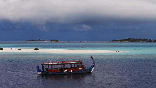 Aerial view of Dhigurah, Maldives shows a dhoni gliding toward a white sandbar and turquoise lagoon. People walk on the sandbank as storm clouds gather over the atoll.
