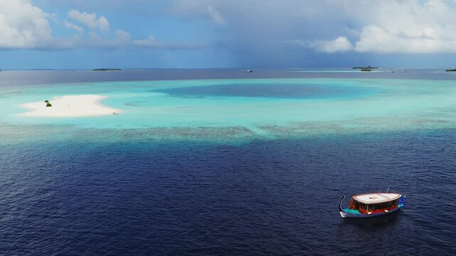Aerial daytime view of Dhigurah island, Maldives. A dhoni drifts, visitors walk a crescent sandbank, coral reef flats encircle, a dark lagoon pool sits, low clouds line the horizon.