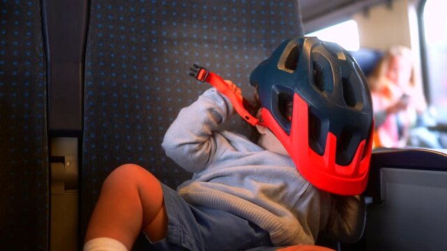 Toddler trying to put on bicycle helmet while riding train with family, playful childhood travel moment highlighting curiosity, independence, exploration and everyday adventure