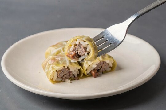Plate of Sarmi with stuffed cabbage rolls filled with minced beef, rice lifted by fork on gray surface. Concept of stuffed cabbage rolls with meat rice in indoor setting.