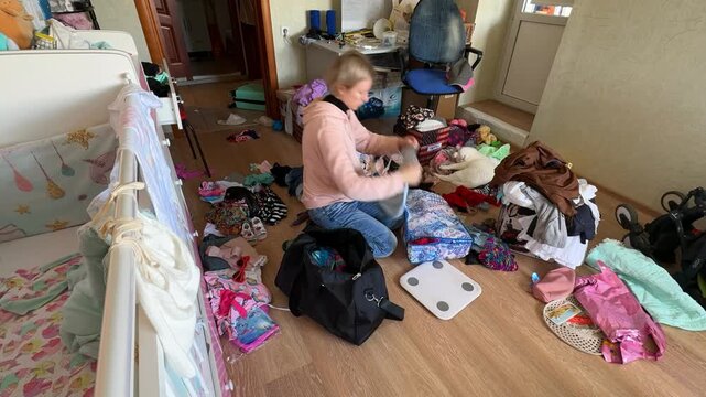 Young woman packing bag in messy nursery. Mother sorting clothes for travel at home. Caucasian girl organizing luggage near baby crib for trip or motherhood plan.