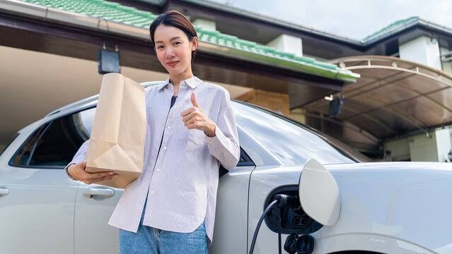 Asian female showing thumbs up sign and holding a paper bag near a charging electric vehicle at home. Concept of easy eco lifestyle and sustainable consumerism.