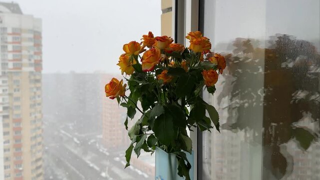 Bouquet of orange roses in vase sitting by window during snowy day. Domestic interior with flowers and city view. Seasonal transition and floral home decoration.