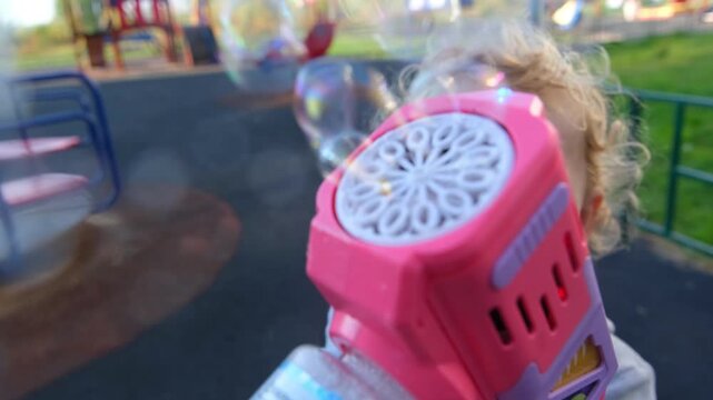 Happy child playing with pink bubble blower toy at playground. Toddler blowing soap bubbles into air outdoors. Childhood activity and fun leisure time concept.