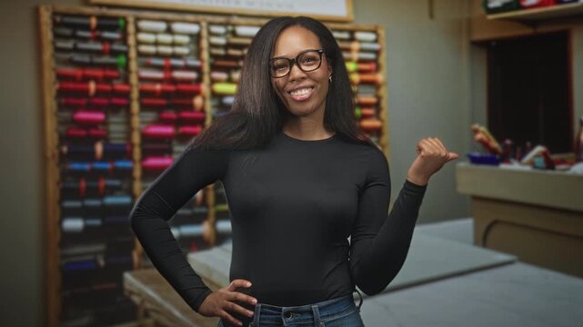Woman with glasses smiles and points thumb to colorful thread shelf in a craft studio; confidence creativity.