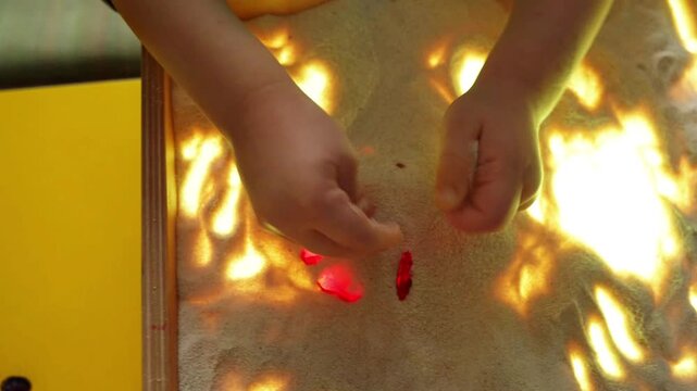Child playing with sand in light box. Kid creating pattern with glass heart on glowing surface. Sensory activity for creativity, learning and child development.