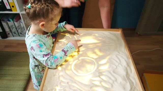 Little girl and woman playing with light sand table. Child learns drawing and sensory therapy. Creative development and education in kindergarten for fun activity.