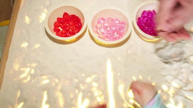Child sorting colored stone gems into bowl on sand table. Educational activity for kid, Montessori sensory game for fine motor skills development, learning concept.