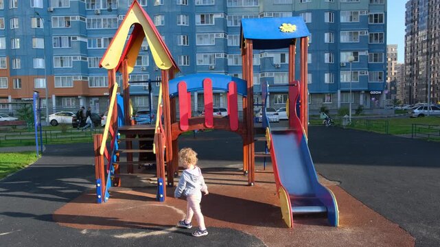 Small kid playing on playground slide outdoors. Toddler girl walking near climbing frame in city courtyard. Childhood activity on modern residential urban area.