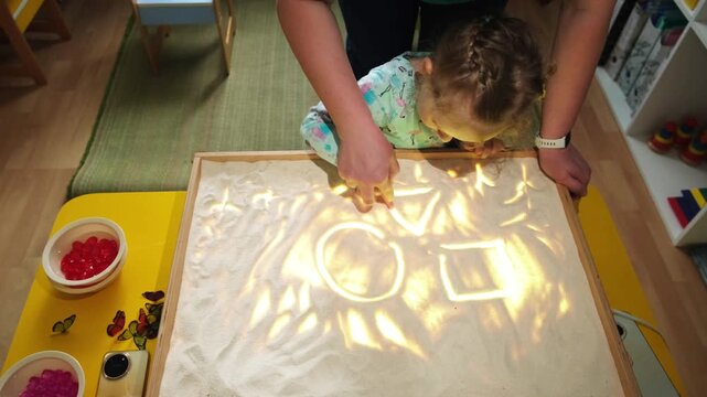 Girl drawing geometric shapes on light box with teacher. Child learning art and geometry using sand on illuminated table. Creative education for preschool kid.