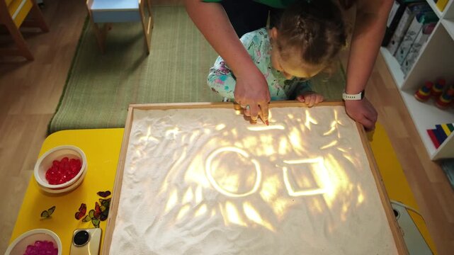 Little girl drawing shapes on a luminous light table with her teacher. Sensory play for development, early education, creative learning and childhood activity.