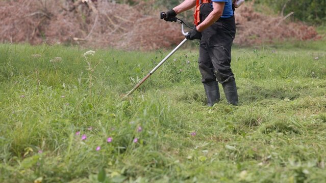 Gardener mowing grass with brushcutter in field next to brushwood pile