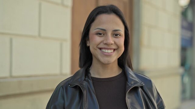 Woman smiling showing teeth with a broad smile, wearing leather jacket and sweater in front of building; joy confidence.