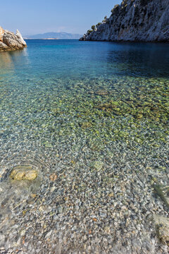 Small pebbles and stones under shallow water by a beach in a cove surrounded by rugged and hilly coastline by the Aegean Sea on Kalymnos island in Greece on a sunny day.