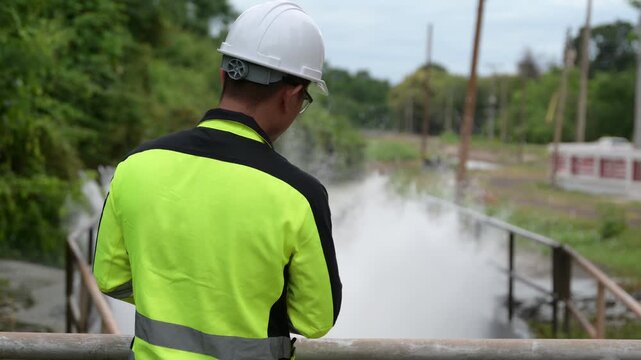 Professional male environmental engineer wearing a hard hat inspecting rushing water at a dam spillway while taking notes on a clipboard for public infrastructure safety and flood control.
