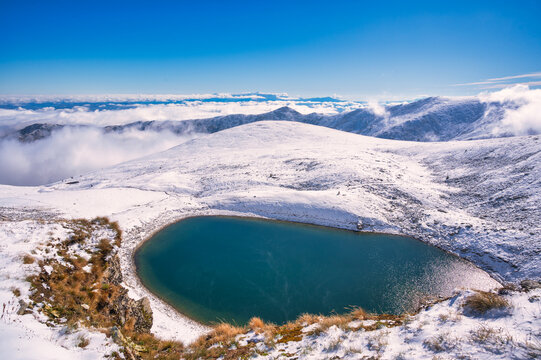 Majestic winter alpine landscape featuring a turquoise glacial lake under a clear blue sky with thick fog rolling over the valley.