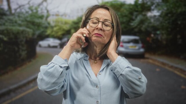 Hispanic woman with grey hair holding smartphone to ear and touching temple on a residential street; concern reflection.