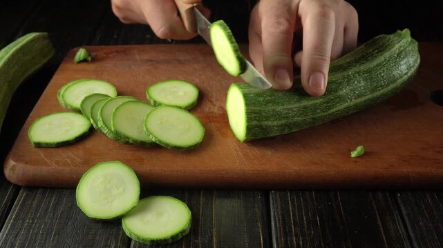 Hands skillfully slice a fresh zucchini into thin rounds on a wooden cutting board, with additional marrow pieces and a dark kitchen surface enhancing the culinary scene