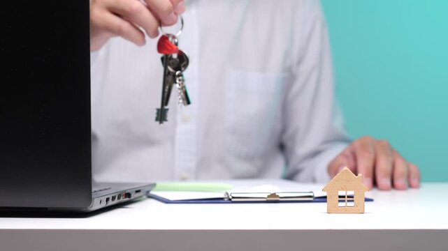 Person in a white shirt displaying a set of house keys, symbolizing successful property acquisition, investment, or rental agreements, with a small wooden house model and contract on the table