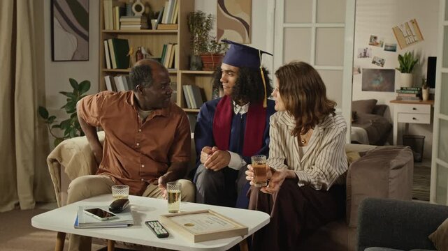 Medium long handheld shot of Black father and Caucasian mother presenting graduation gift to young adult son wearing academic cap. Video suitable for student loan or university marketing ads