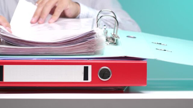 Person's hands organizing numerous paper documents inside a crowded blue ring binder. Stacked upon a red folder on a light desk against a blue background
