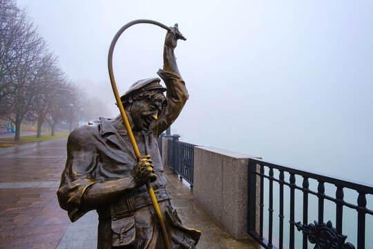 Rostov-on-Don, Russia - November 28, 2019: Close-up of bronze fisherman statue holding a net pole on foggy riverside promenade with wet pavement