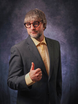Lawyer portrait with thumbs up gesture. Man in his 40s or 50s wearing dark grey suit and yellow shirt, Male with grey beard and hair and glasses, dark painted background. Calm and confident expert