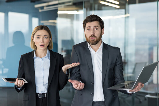 Two business coworkers standing in a modern office, one holding a tablet and the other a laptop, expressing confusion and doubt with open hands and shrugging shoulders