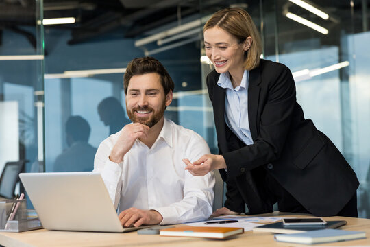 Business colleagues smiling while collaborating on a project, discussing new strategy and solutions using a laptop at their desk in a contemporary corporate office environment