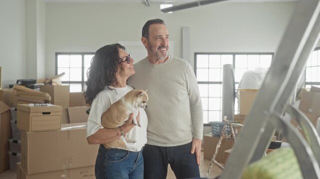 Woman holding small dog, man with arm around her amid moving boxes in a building; contentment new home.