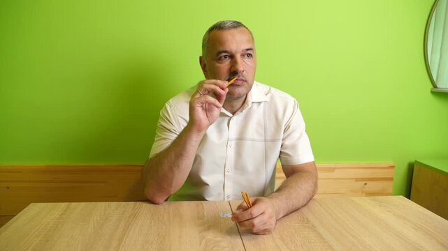 A man sits at a table eating a breadstick snack while quietly enjoying a light snack in a bright room with green walls.