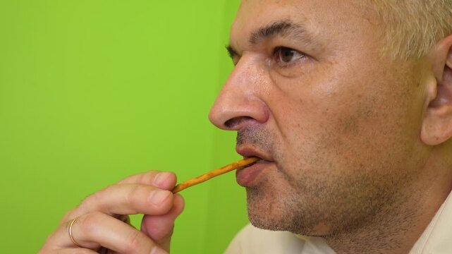 Close up of a man eating a crunchy breadstick snack while slowly snacking and looking to the side in a bright room.