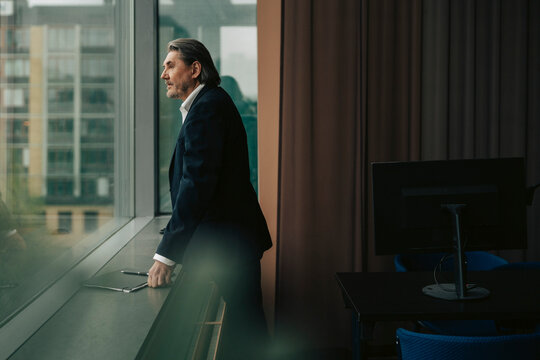 Contemplative senior male corporate professional looking out through window while standing near sill in office