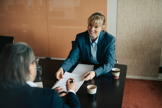 Smiling female finance advisor passing documents to coworker sitting at table