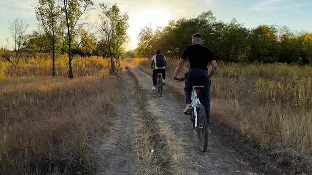 Young male friends ride mountain ski bikes along trail on sunny summer day. Beautiful golden sunlight at background. Concept of active lifestyle, adventure spirit and carefree weekend outdoor