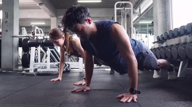 Asian man and woman performing push-ups side by side on a black rubber mat inside a fitness center. Professional and motivating for bodybuilding, strength training, and healthy lifestyle concepts.