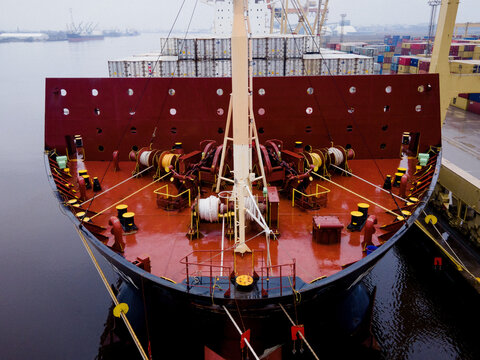 Aerial view of the bow of a large container ship docked at the port with mooring winches, ropes, and colorful shipping containers in the background Riga, Latvia.