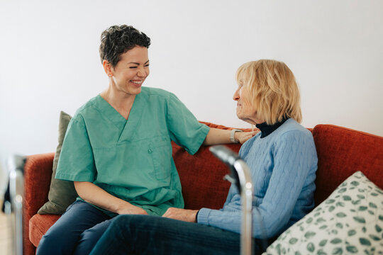 Happy female caregiver sitting on couch and talking with senior woman at nursing home
