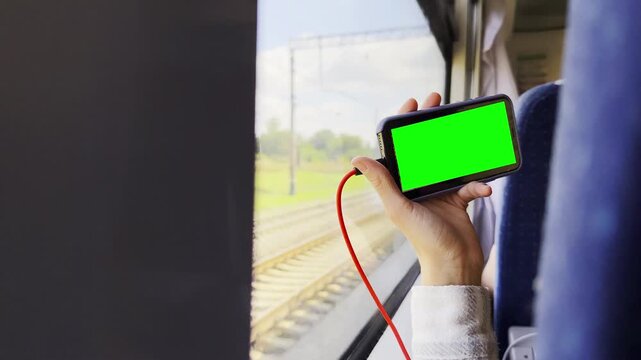 Close up of female hand holding smartphone with green screen while traveling by train. Young woman watching videos during journey on passenger railway transport. The scene conveys relaxation on road