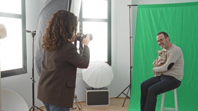 Man sits on stool holding small dog while woman photographer aims camera in studio; friendly warmth.