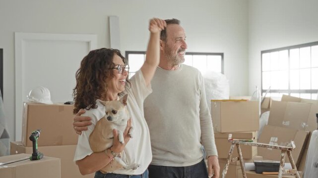Man with arm around woman holding small dog among stacked moving boxes, ladder and drill in a bright building; contentment new home.