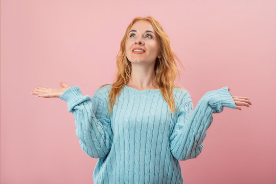 Smiling woman with reddish hair and blue eyes looking upwards, holding both hands open, palms facing up, against a pink background