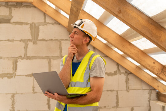 Construction worker wearing a hard hat and safety vest, holding a laptop and contemplating plans in an unfinished building with wooden roof beams and brick walls
