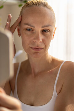 Woman closely examining her reflection in a mirror, carefully checking skin for signs of aging, wrinkles, and fine lines while performing her daily skincare routine in the morning light