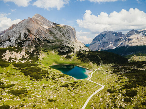 Aerial view of Lago Limo surrounded by lush green alpine meadows, rocky mountain peaks, and a winding trail under a clear blue sky in Marebbe, Trentino-Alto Adige, Italy.