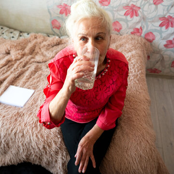 Elderly woman enjoys refreshing water while sitting comfortably on a plush bed adorned with soft textures and vibrant floral patterns in a cozy indoor setting